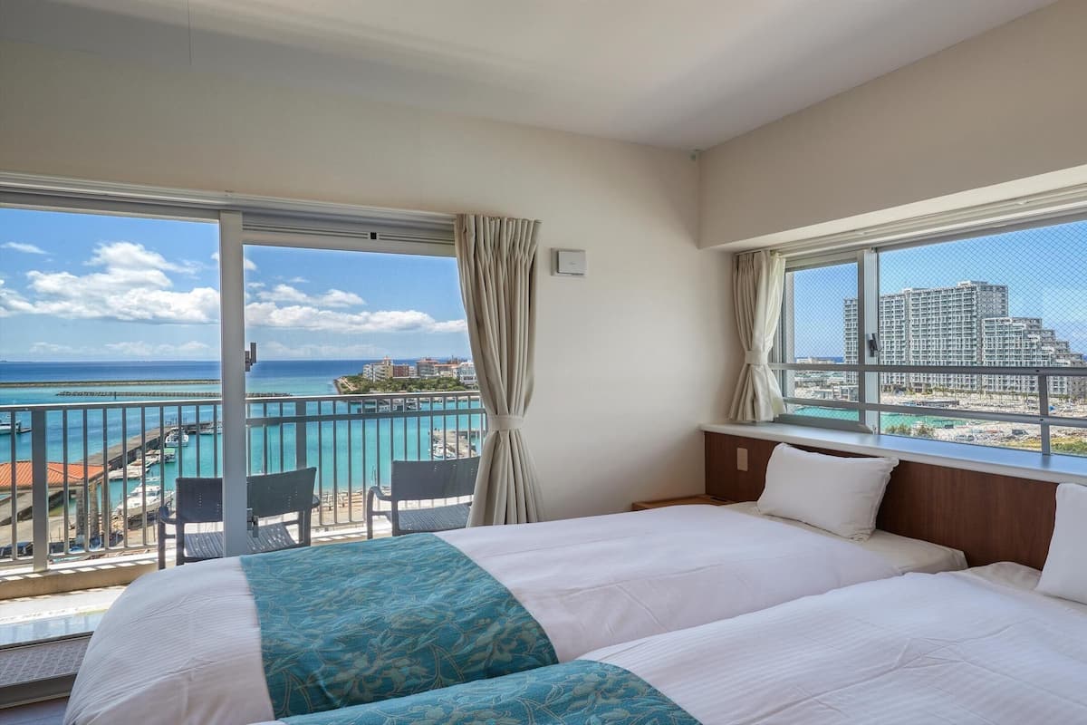 A hotel room with two beds, a balcony with a view of the sea and buildings, and a window with a view of the cityscape.