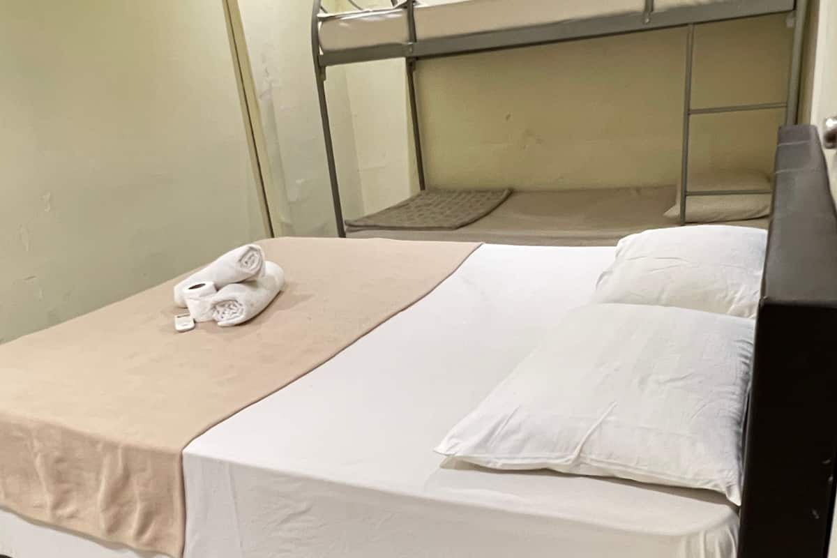 A single bed with a beige bedspread and white pillows, a metal bunk bed in the background, and a small towel on the bed.