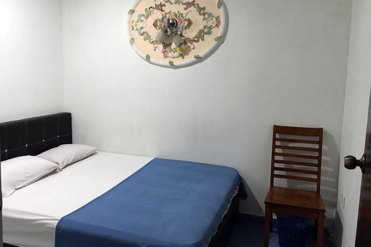 A bedroom with a bed, a blue bedspread, a wooden chair, and a decorative wall hanging.