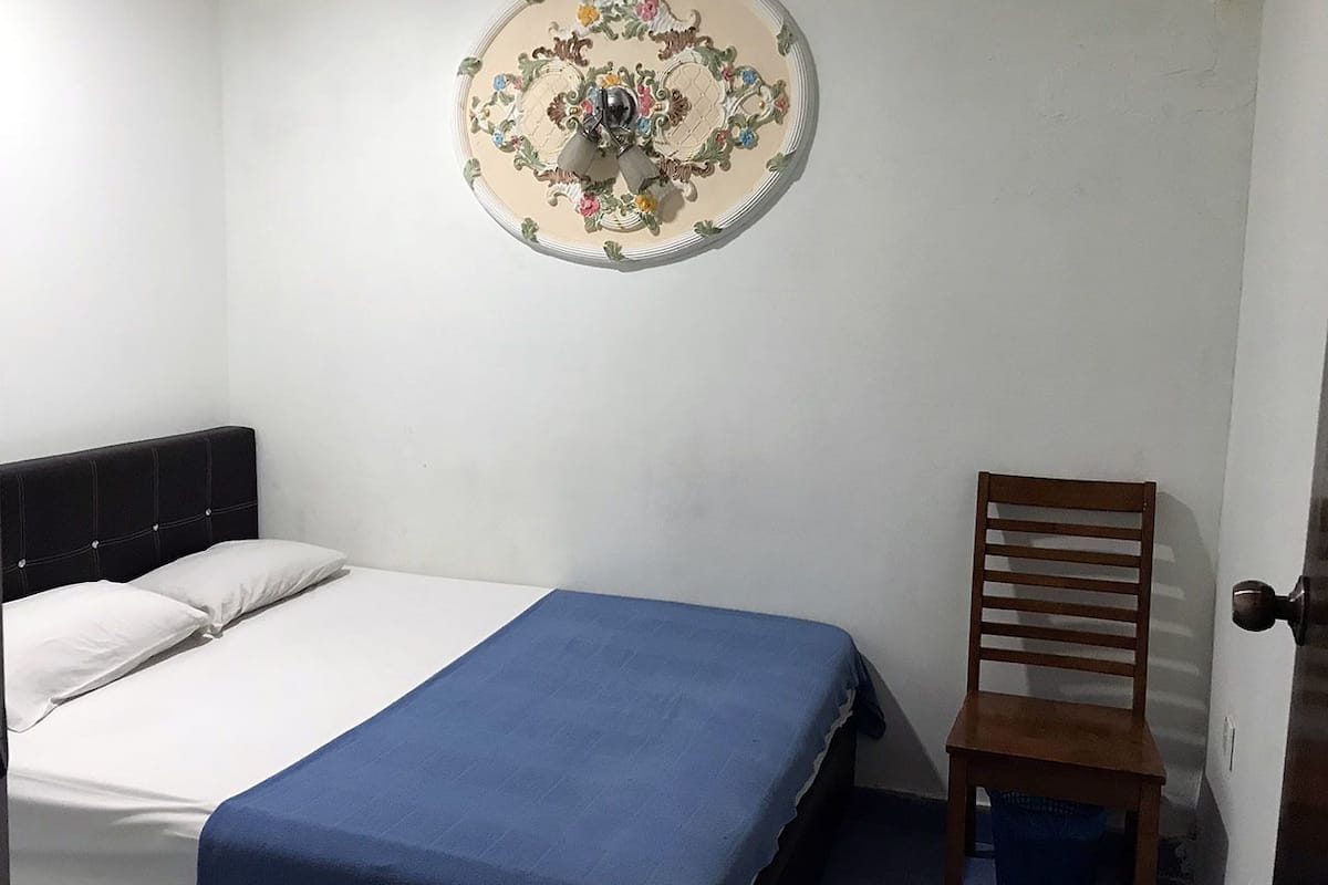 A room with a bed, a blue bedspread, a wooden chair, and a decorative wall hanging.