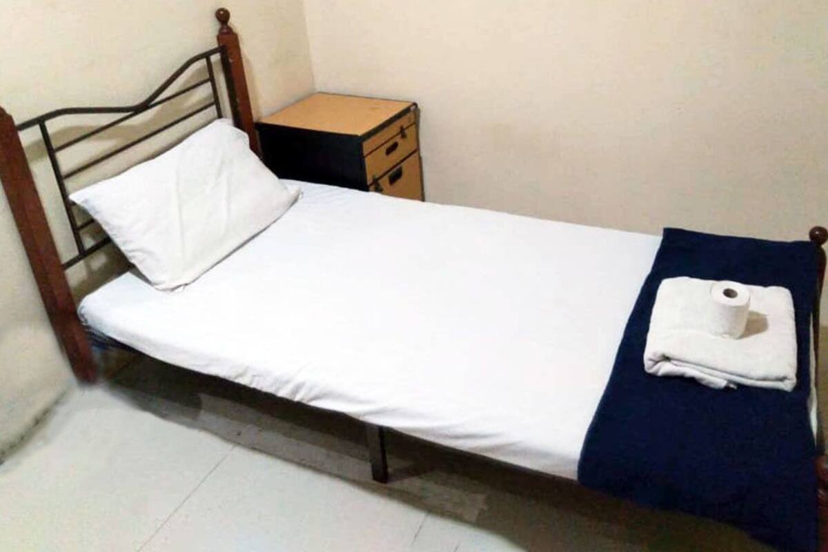 A single bed with a wooden frame, white bedding, and a blue blanket. There is a small wooden nightstand with a drawer and a roll of paper towels.