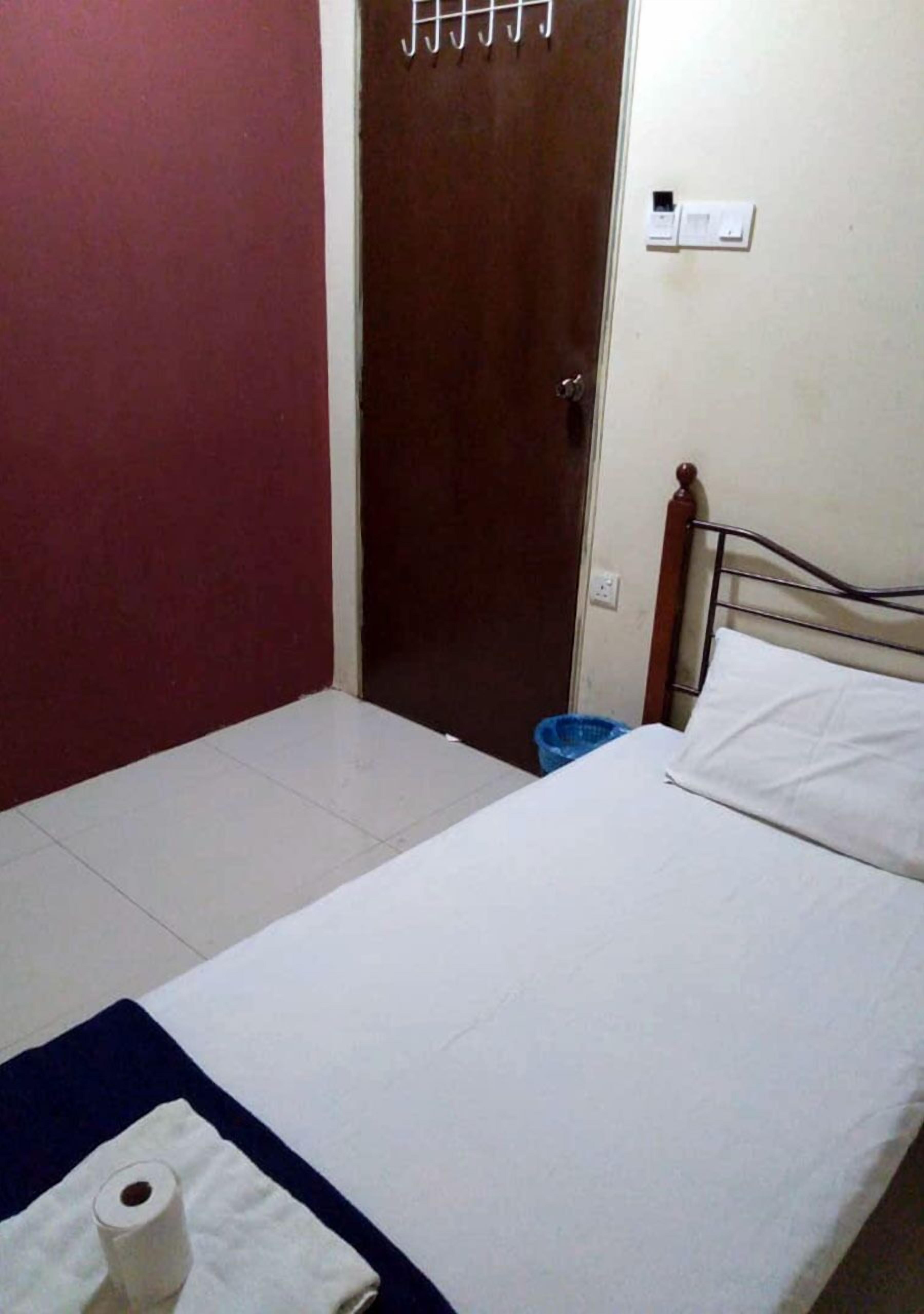 A small, simple room with a bed, a wooden door, a blue trash bin, and a white wall with a light switch.