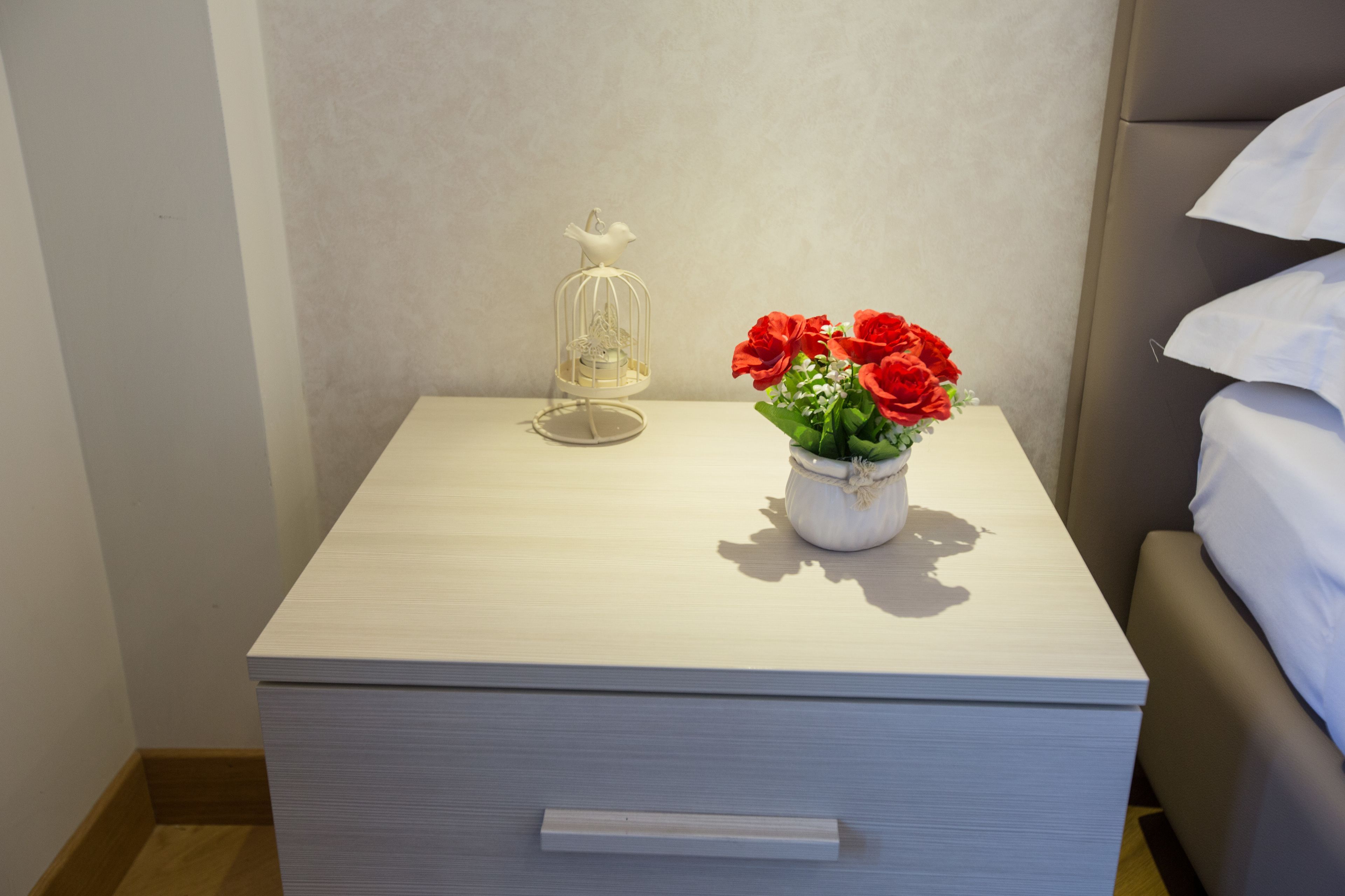 A neatly made bed with white linens, a bedside table with a white vase of red flowers, and a white birdcage with a small item inside.