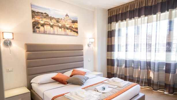 A hotel room with a large bed, a nightstand, a wall-mounted lamp, and a window with curtains. - Vatican City Relais (Rome)