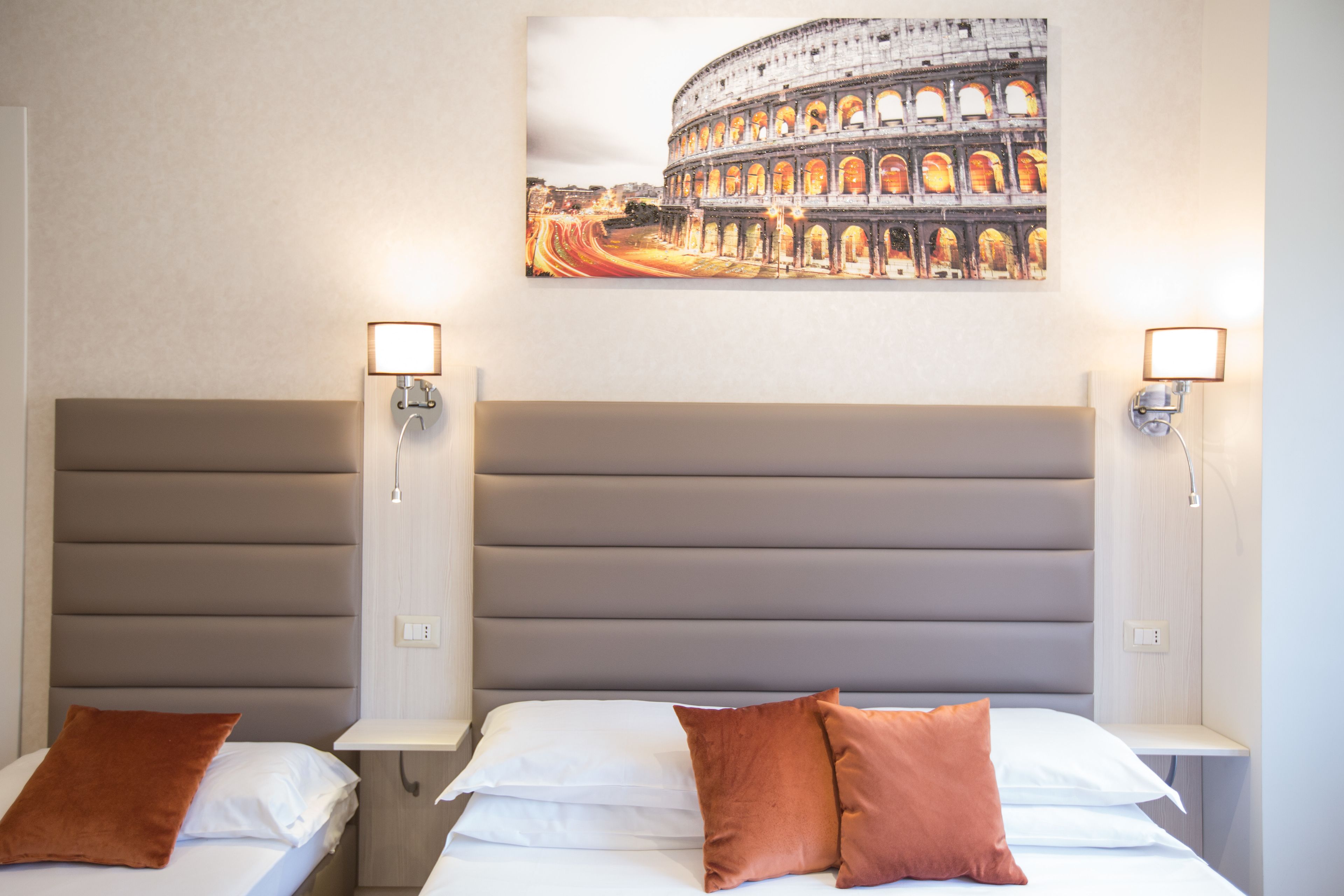 Habitación de hotel con dos camas, cada una con un almohadón marrón, un cabecero grande y una fotografía enmarcada del Coliseo en la pared.