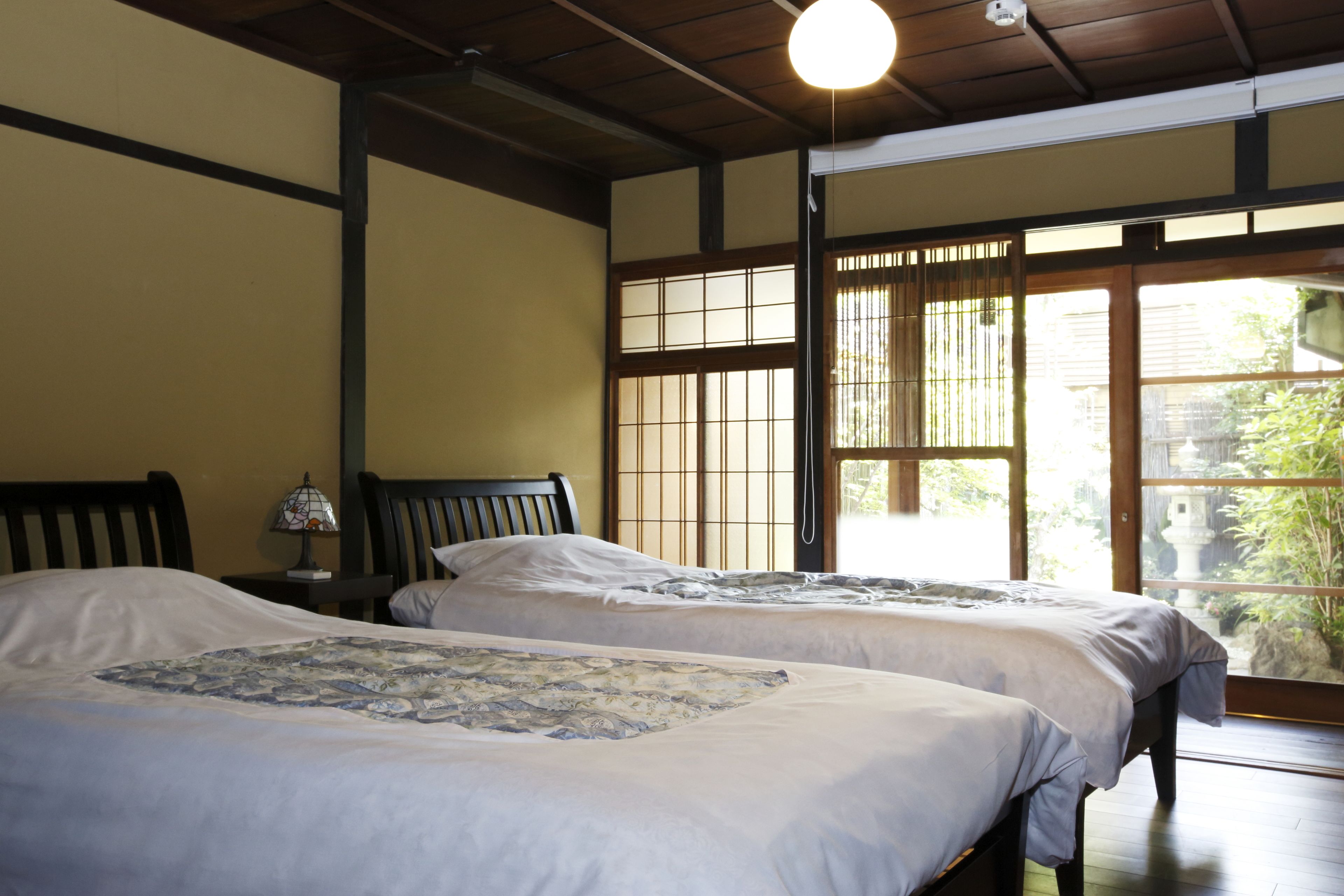 A traditional Japanese-style room with two beds, a wooden ceiling, and a view of greenery outside.