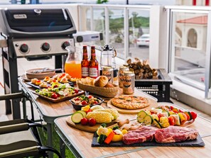 Un coin barbecue avec divers plats et boissons disposés sur une table, un gril en arrière-plan et une vue sur la rue à travers une fenêtre.