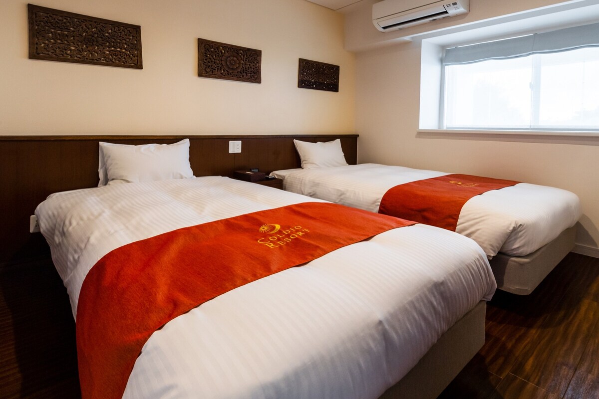 Habitación de hotel con dos camas, cada una con ropa blanca y colchas anaranjadas. Se observan paneles decorativos en la pared sobre las camas y una ventana con vistas.
