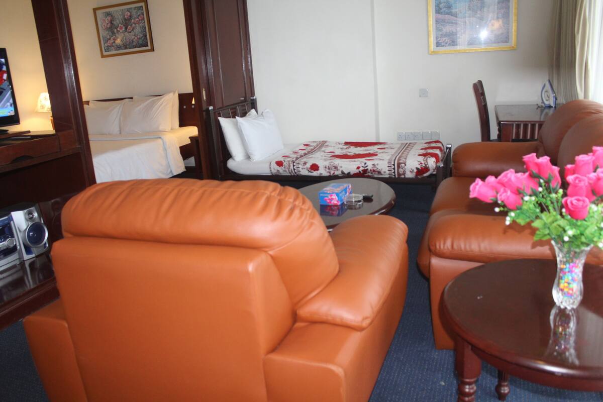 A hotel room with a brown leather sofa, a small round table with a vase of pink flowers, a bed with a floral bedspread, and a television on a wooden stand.