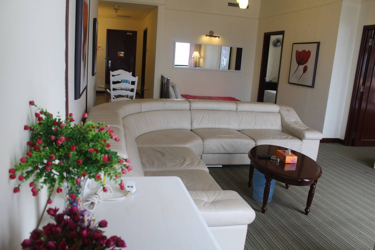 A living room with a beige sectional sofa, a white coffee table, and a small side table with a vase of flowers. There is a mirror on the wall and a framed picture above it.