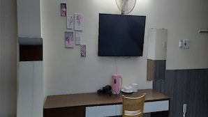 A hotel room with a desk, a chair, a flat-screen TV mounted on the wall, a small cabinet, and a few framed pictures on the wall. - Ailen Garden II Hotel (Ho Chi Minh City)