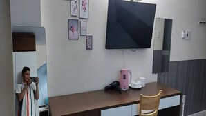 A modern hotel room with a large flat-screen TV mounted on the wall, a wooden desk with a chair, a pink electric kettle, and a small table with cups. - Ailen Garden II Hotel (Ho Chi Minh City)