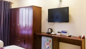 A hotel room with a wooden wardrobe, a flat-screen TV mounted on the wall, a small refrigerator, a desk with a kettle, cups, and bottled water. - Ailen Garden II Hotel (Ho Chi Minh City)