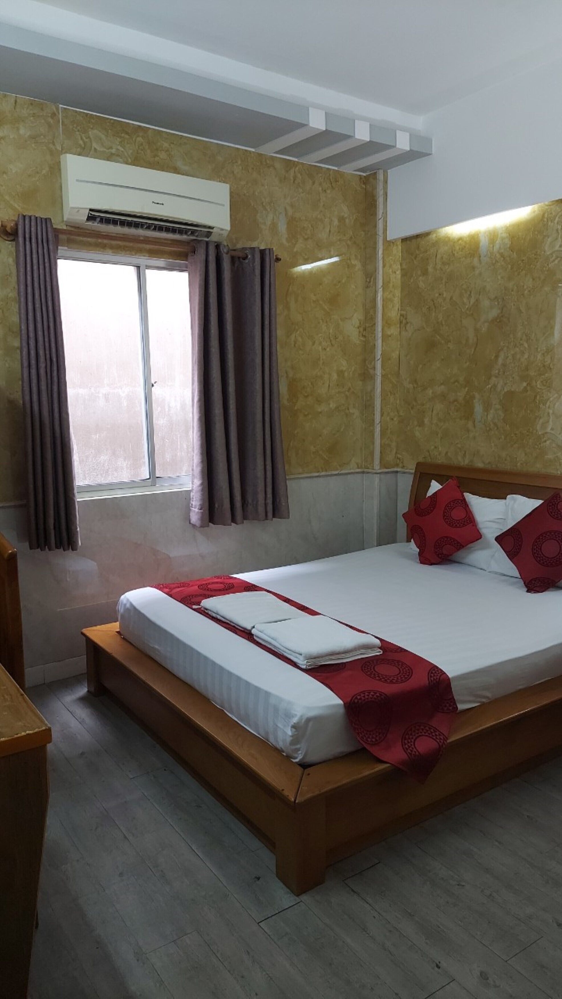Habitación de hotel con cama, cabecera de madera, ventana con cortinas, equipo de aire acondicionado y piso de madera.