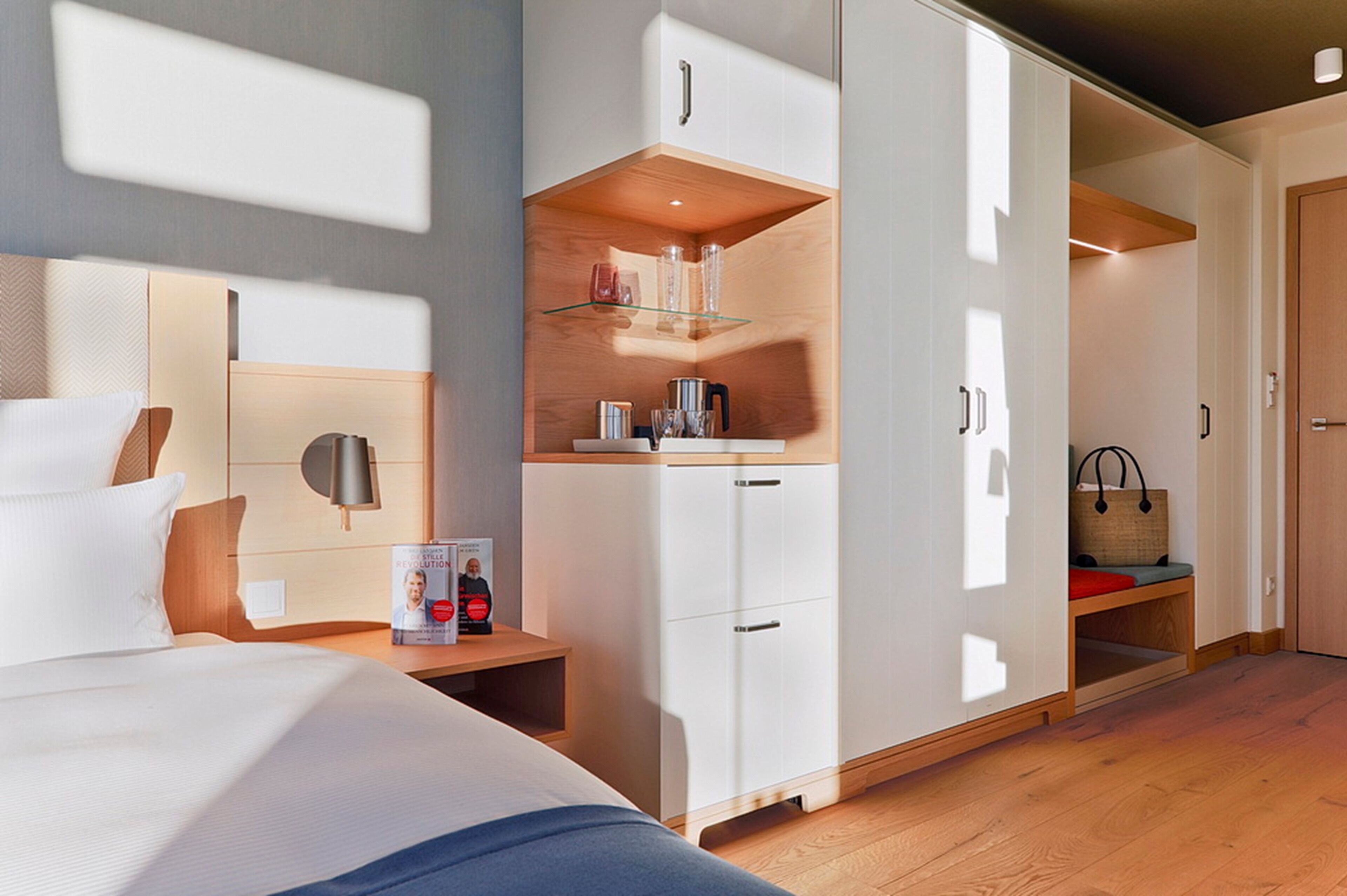 Una habitación de hotel moderna con cama, una mesita de noche con una revista, un armario con repisa de vidrio y un banco de madera.