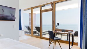 A hotel room with a large sliding glass door leading to a balcony with a view of the sea. There is a bed, a desk with a chair, and a TV mounted on the wall. - Upstalsboom Wyk auf Föhr (Wyk auf Foehr)