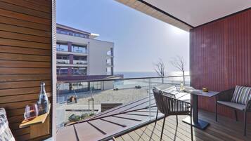 Een balkon met een houten vloer, een tafel met een fles en glazen, en uitzicht op een gebouw en strand.