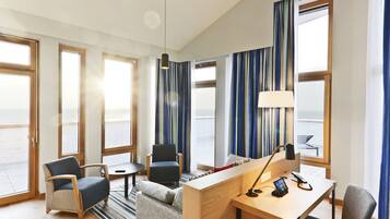 Une chambre d’hôtel équipée d’un bureau, de deux fauteuils, d’un canapé, d’une petite table, d’une lampe et de grandes fenêtres avec des rideaux.