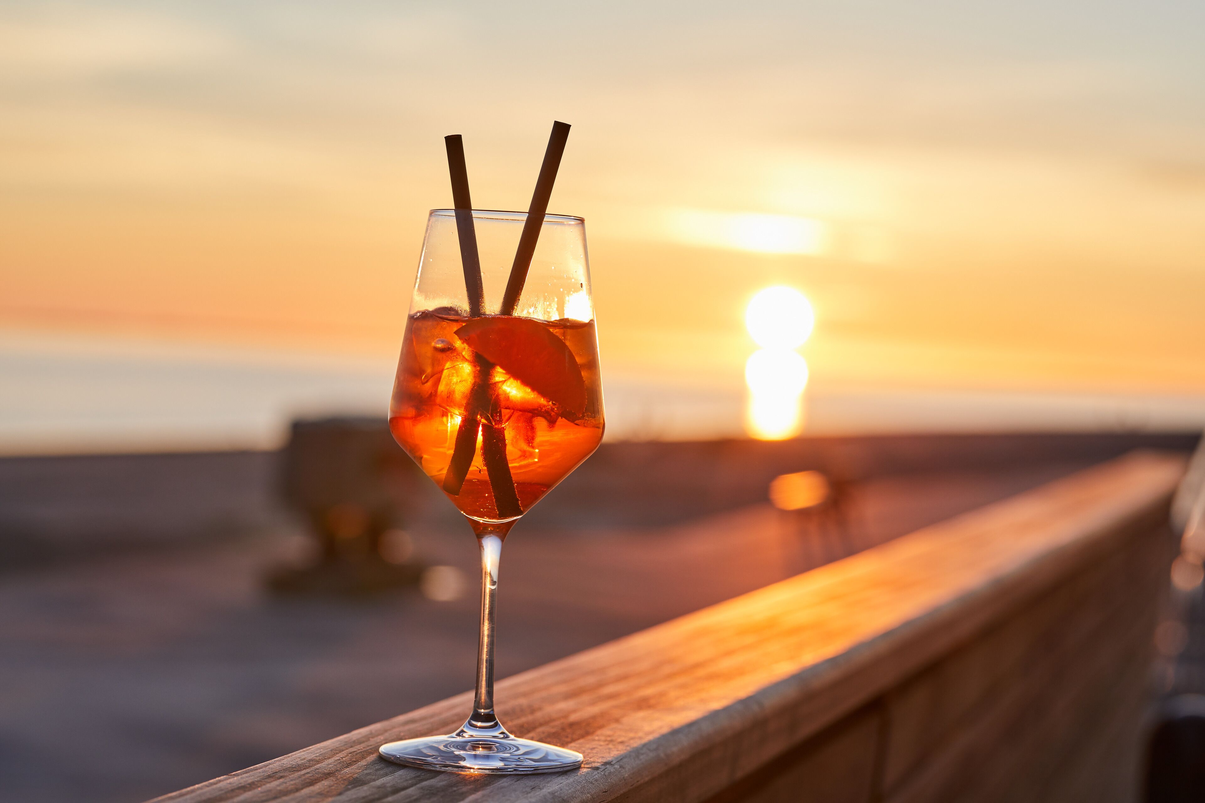 Cocktail-lasi, jossa on sitruunaviipale ja kaksi pilliä, asetettu puiseen kaiteeseen, ja taustalla näkyy auringonlasku.
