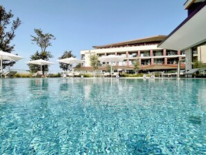 Una piscina cristalina con sillones de descanso y sombrillas, un edificio moderno con un letrero que dice "Upstateroom.com", y un cielo azul despejado.