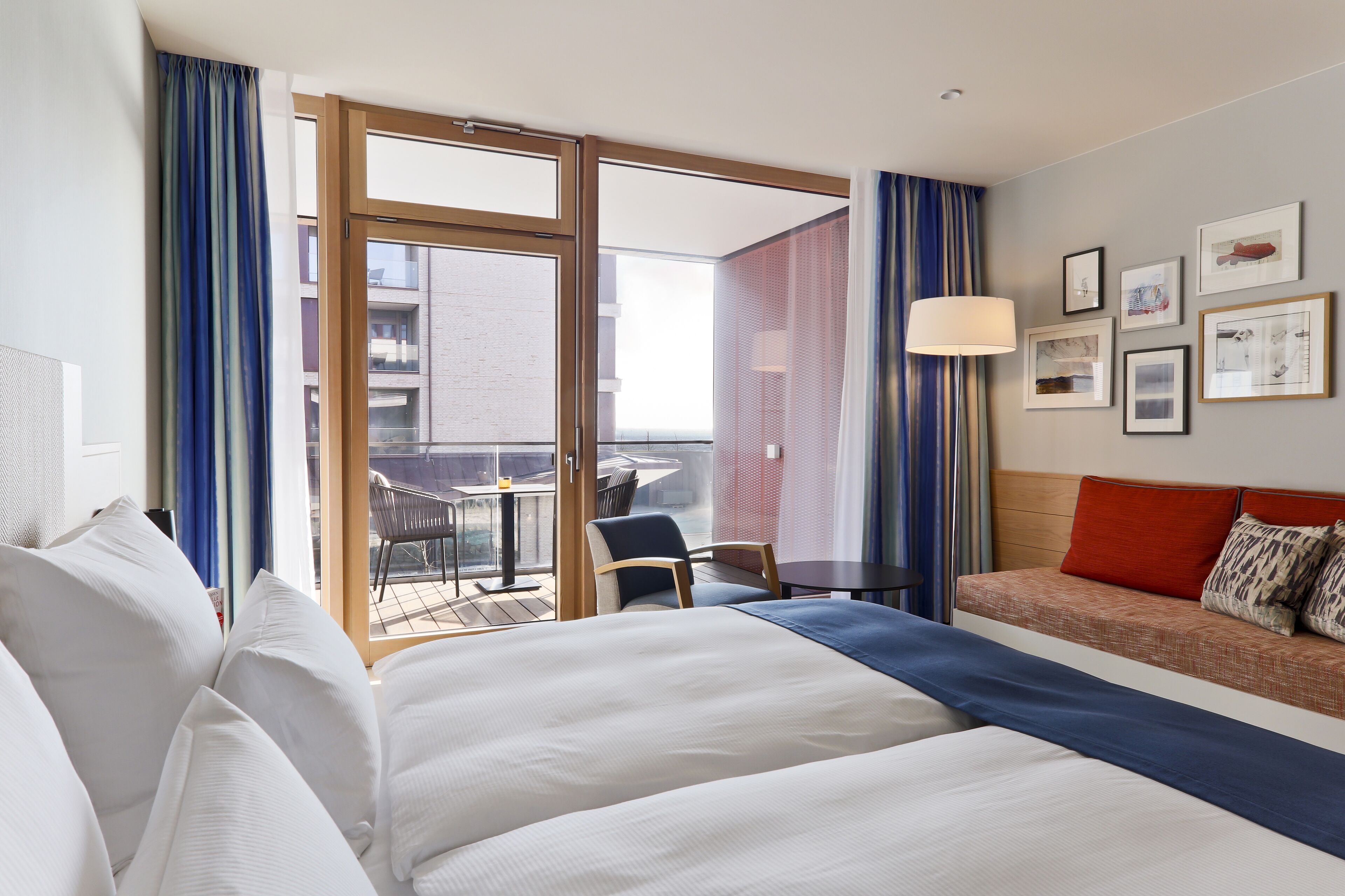 A hotel room with a large bed, a sofa, a balcony with a table and chairs, and framed pictures on the wall.