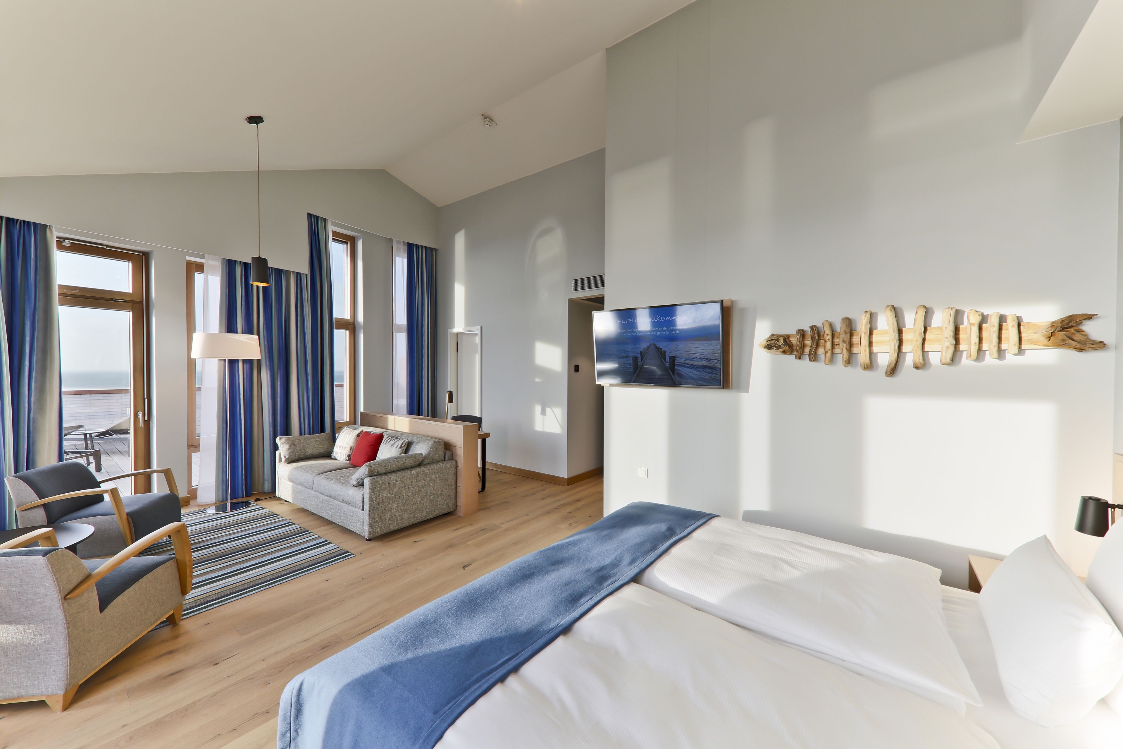 A modern hotel room with a large bed, a sofa, a desk, and a view of the sea through the windows.