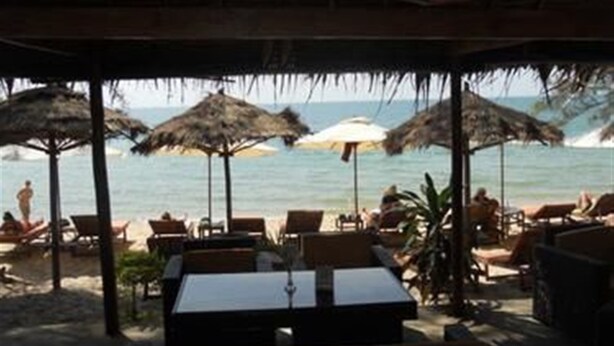 An outdoor seating area with tables and chairs under a thatched roof, overlooking a beach with umbrellas and people. - Chez Paou (Sihanoukville)