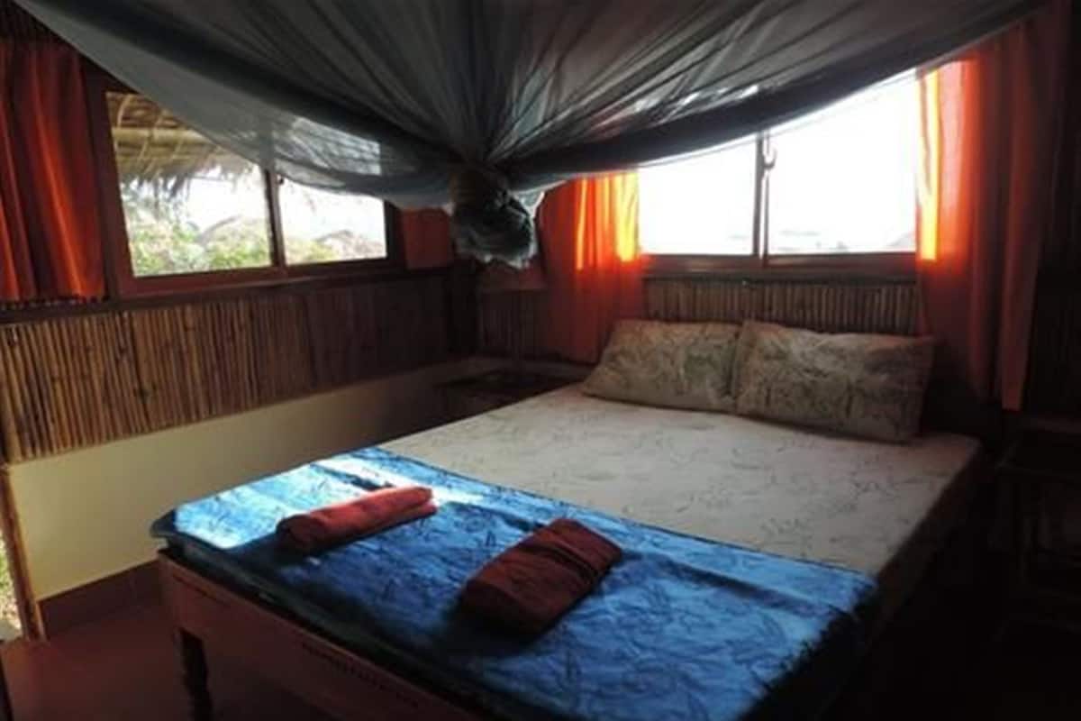 A single bed with a blue bedspread and two red pillows, a wooden headboard, and a window with curtains.
