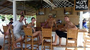 A group of people seated at a table under a covered area with a sign reading "HOT DOGS". - Chez Paou (Sihanoukville)