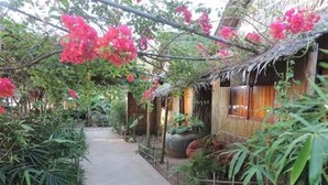 A pathway leading to a rustic building with a thatched roof, surrounded by lush greenery and flowering plants. - Chez Paou (Sihanoukville)