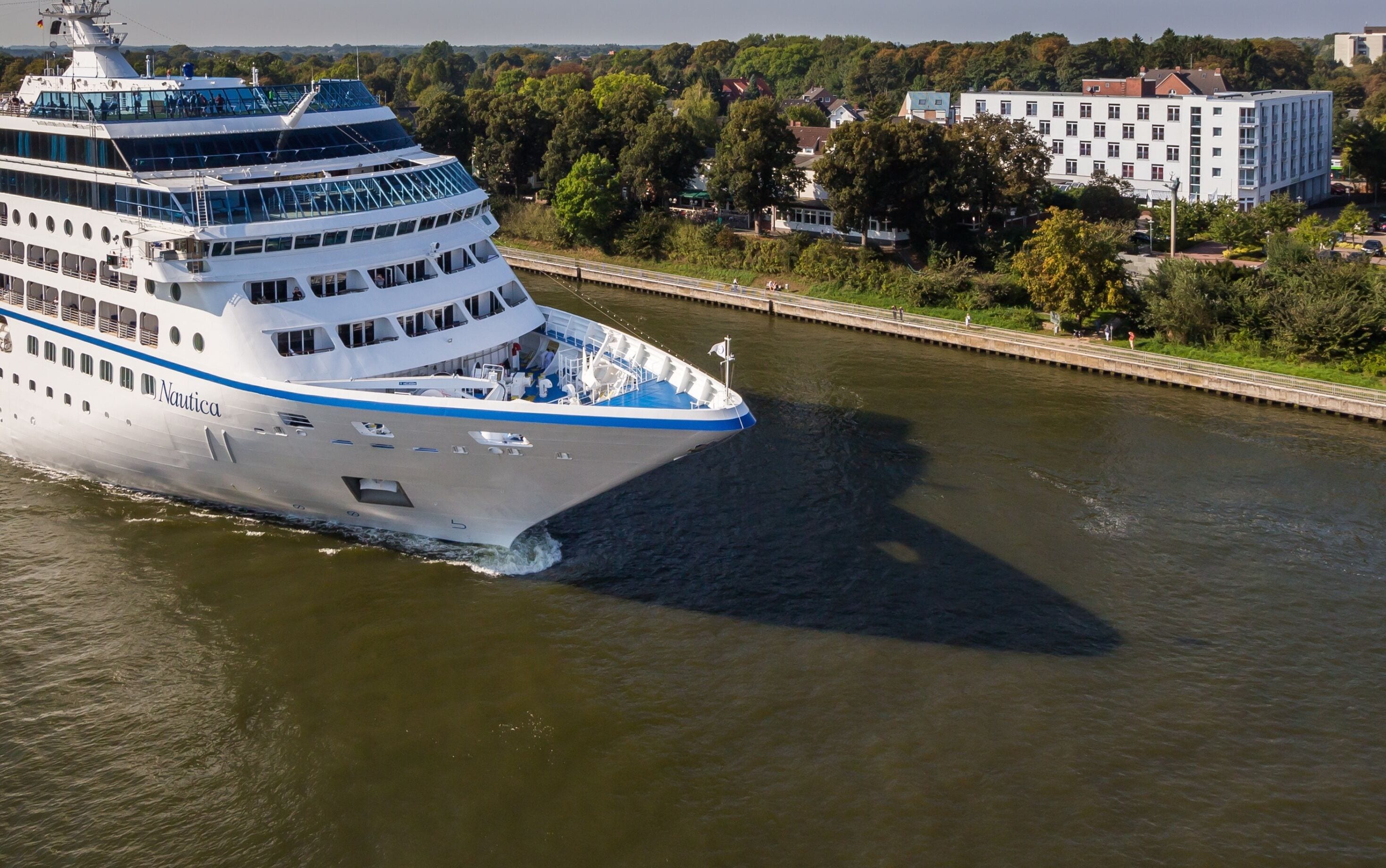 Een groot cruiseschip met meerdere dekken en een blauwe streep, afgemeerd in een haven met een brug en gebouwen op de achtergrond.