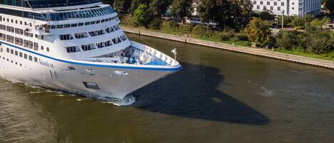 Un gran crucero con varias cubiertas y una franja azul, amarrado en un puerto con un puente y edificios al fondo.
