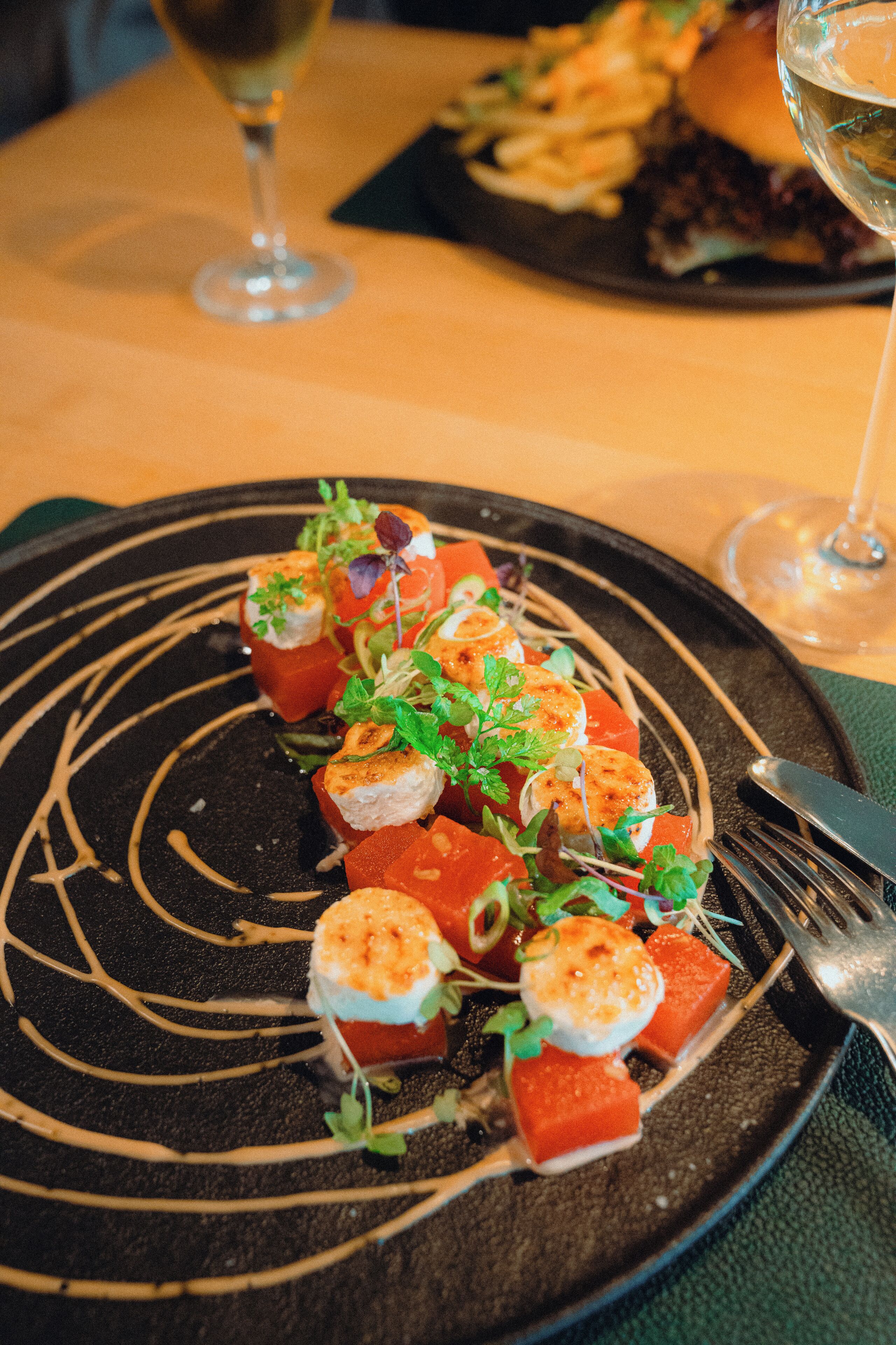 A plate of food with a black rim and white sauce, a fork, and a glass of white wine on a table.