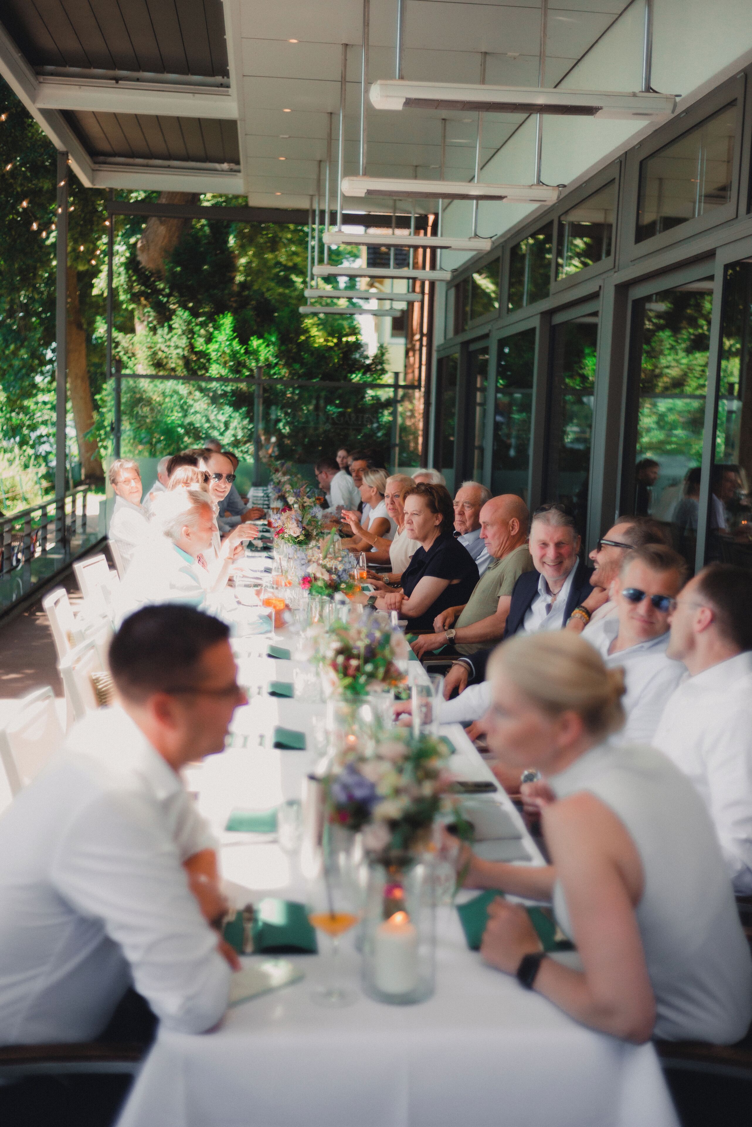 Una larga mesa de comedor con invitados sentados, rodeada de ventanales y follaje.