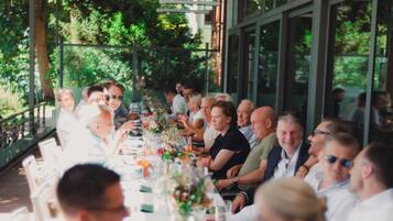 Una larga mesa de comedor con invitados sentados, rodeada de ventanales y follaje.