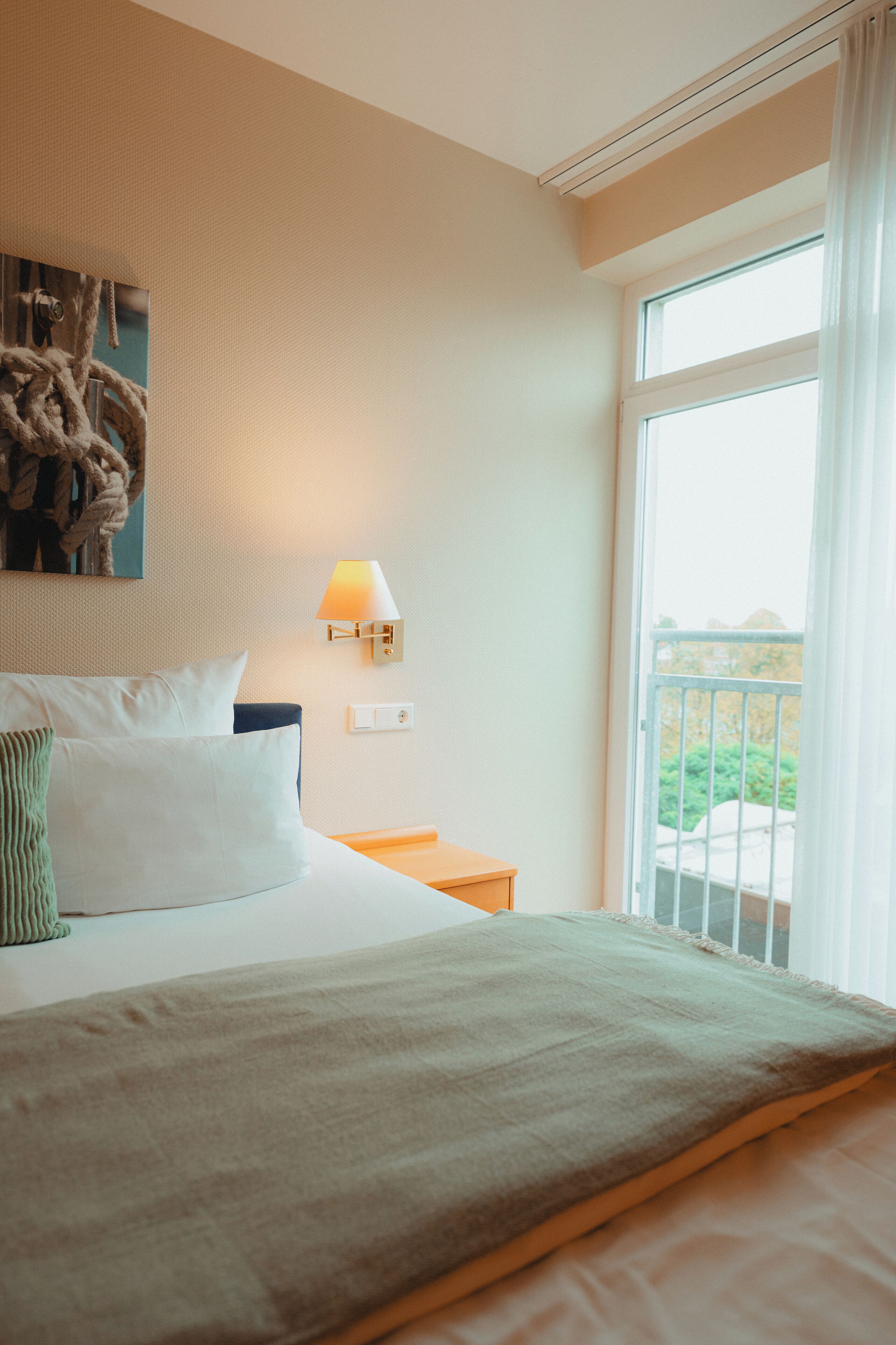 Habitación de hotel con cama, mesita de noche, lámpara de pared y ventana con vista a la vegetación.