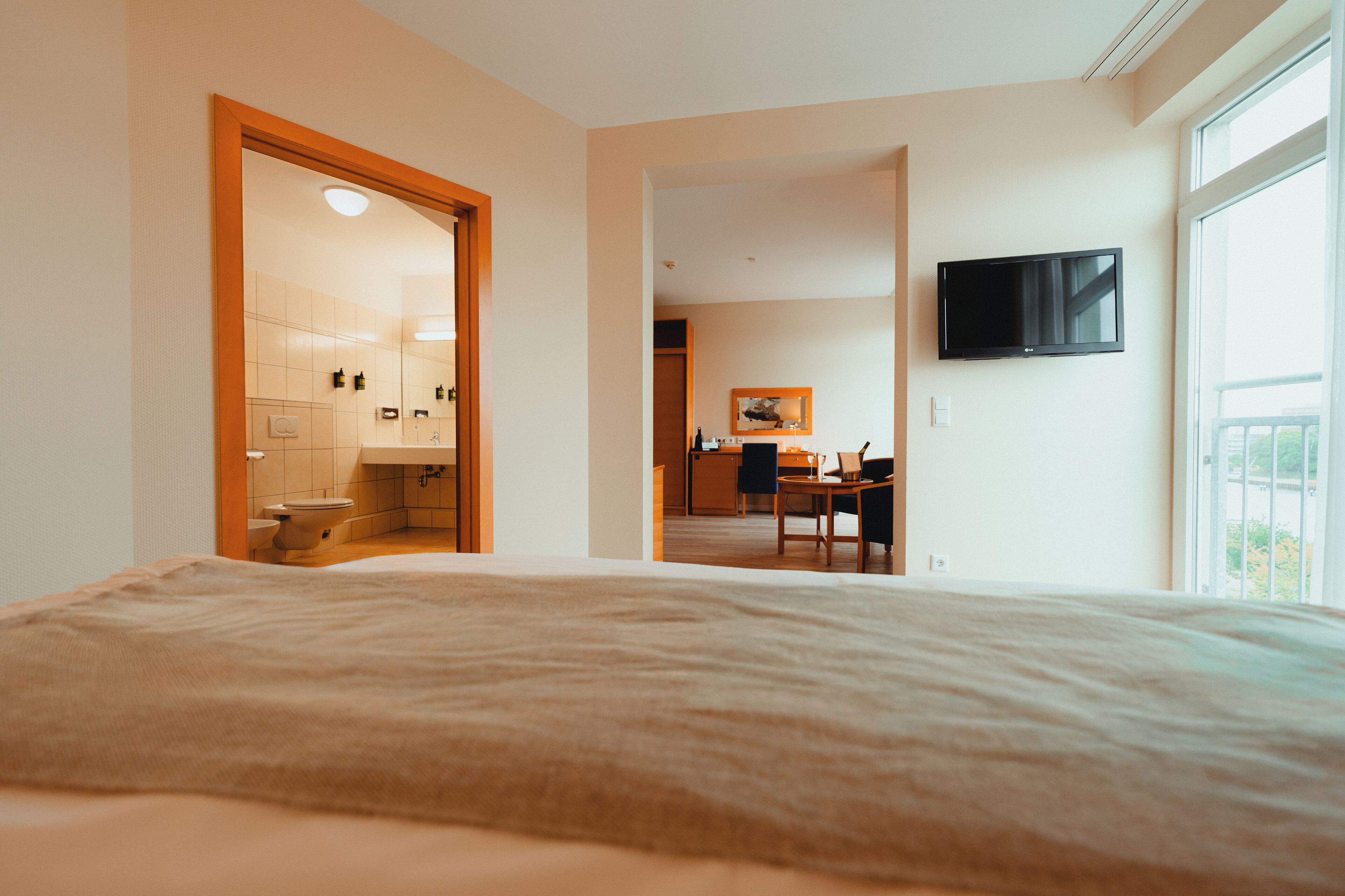 A hotel room with a bed, a TV mounted on the wall, and a view of the bathroom and kitchen area.