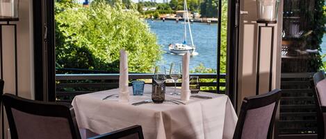 A dining table set with candles, glasses, and cutlery, overlooking a river with boats and a bridge.