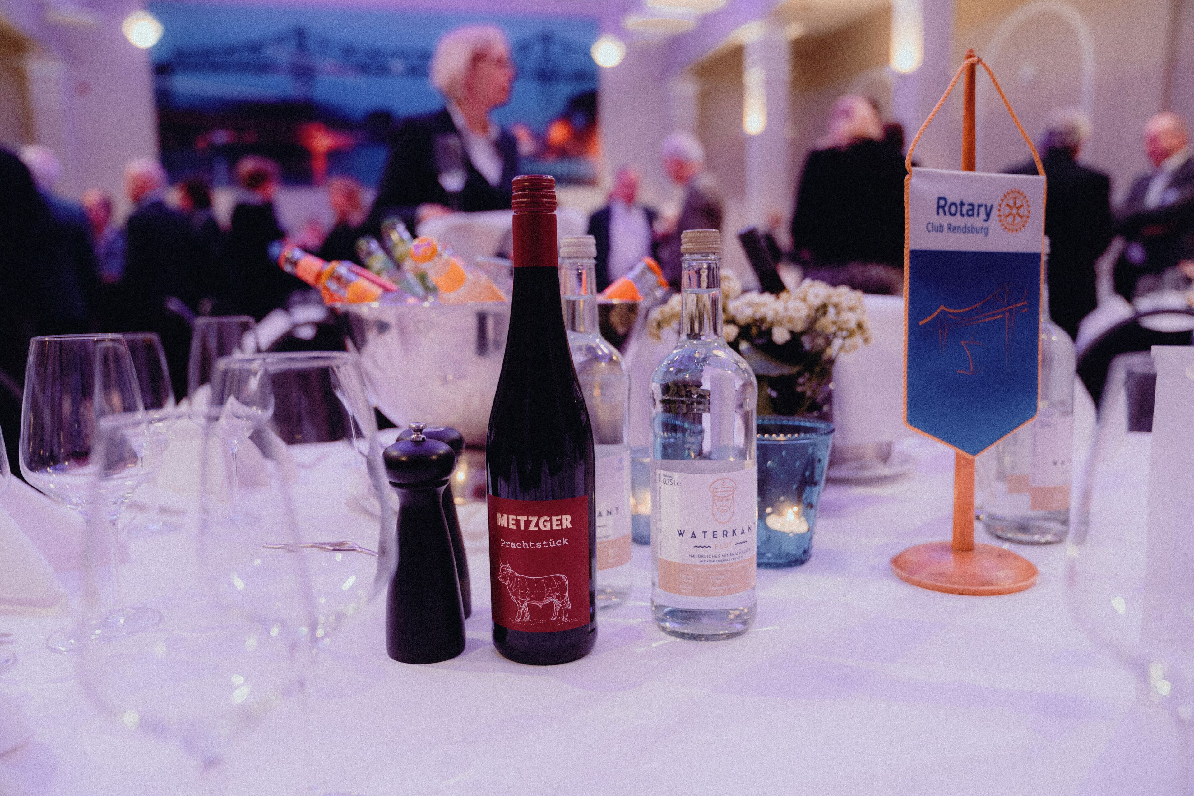 Mesa dispuesta con botellas de vino, copas y una botella de agua, con un cartel del Club Rotary al fondo.