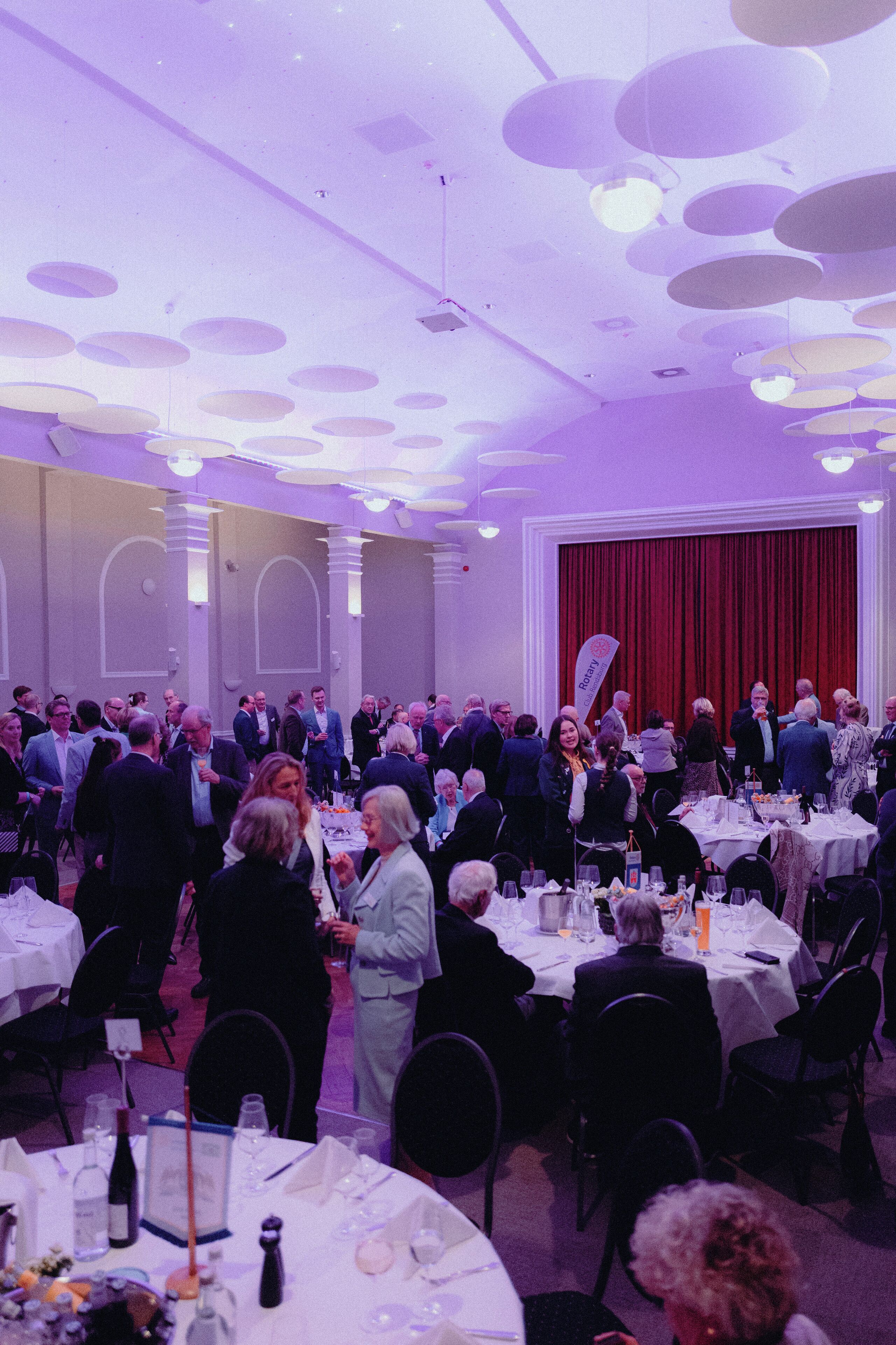 A formal event with round tables, attendees in business attire, and a stage with curtains in the background.