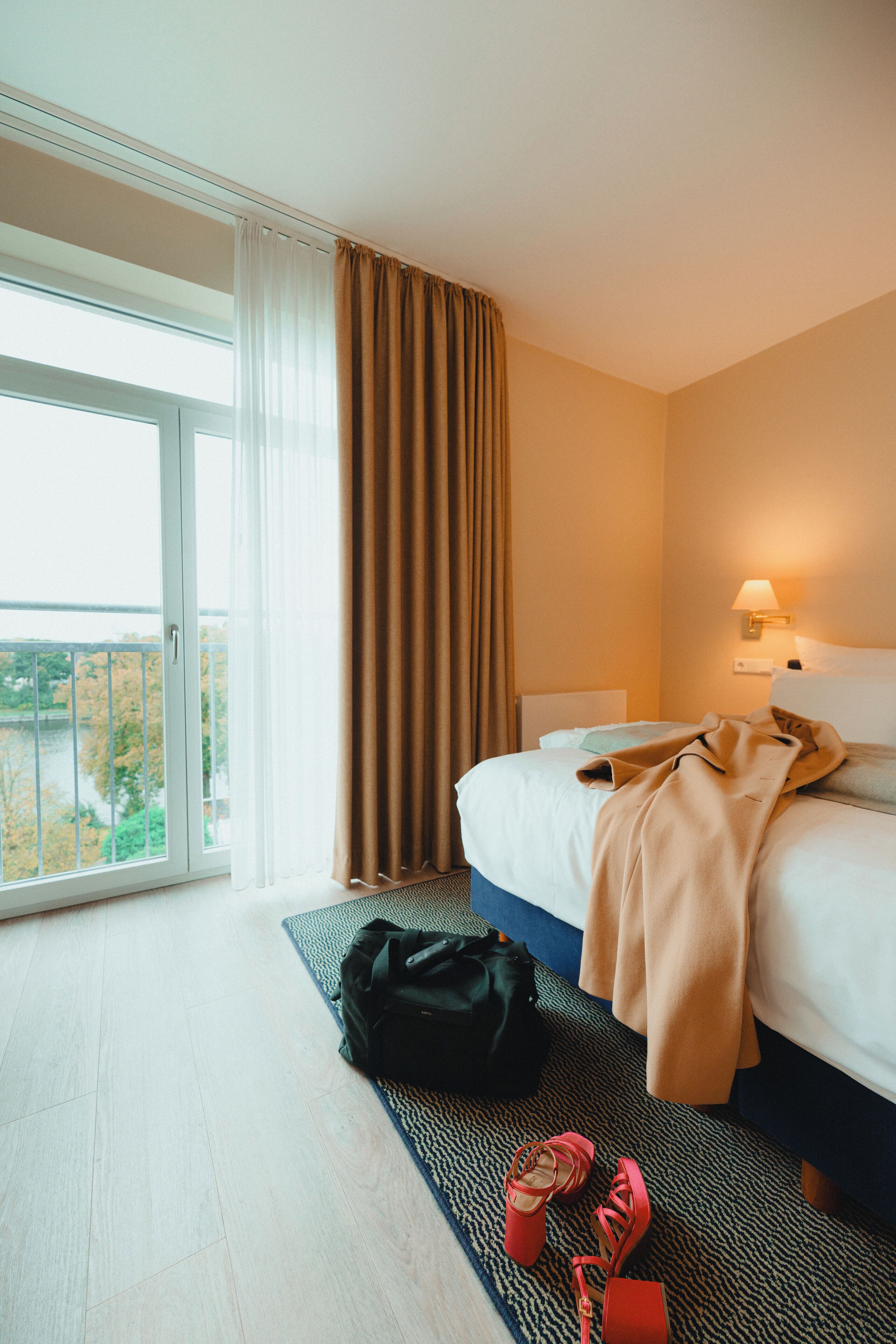 Una habitación de hotel con cama, mesita de noche, lámpara, ventana con cortinas y vistas al exterior.