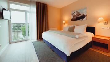 A hotel room with a large bed, a flat-screen TV mounted on the wall, a painting above the bed, and a window with curtains.
