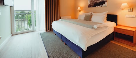 A hotel room with a large bed, a flat-screen TV mounted on the wall, a painting above the bed, and a window with curtains.