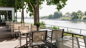 Un espacio de comedor al aire libre con mesas y sillas, con vista a un río y un puente al fondo.
