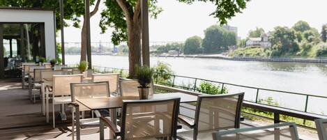 An outdoor dining area with tables and chairs, overlooking a river with a bridge in the background.