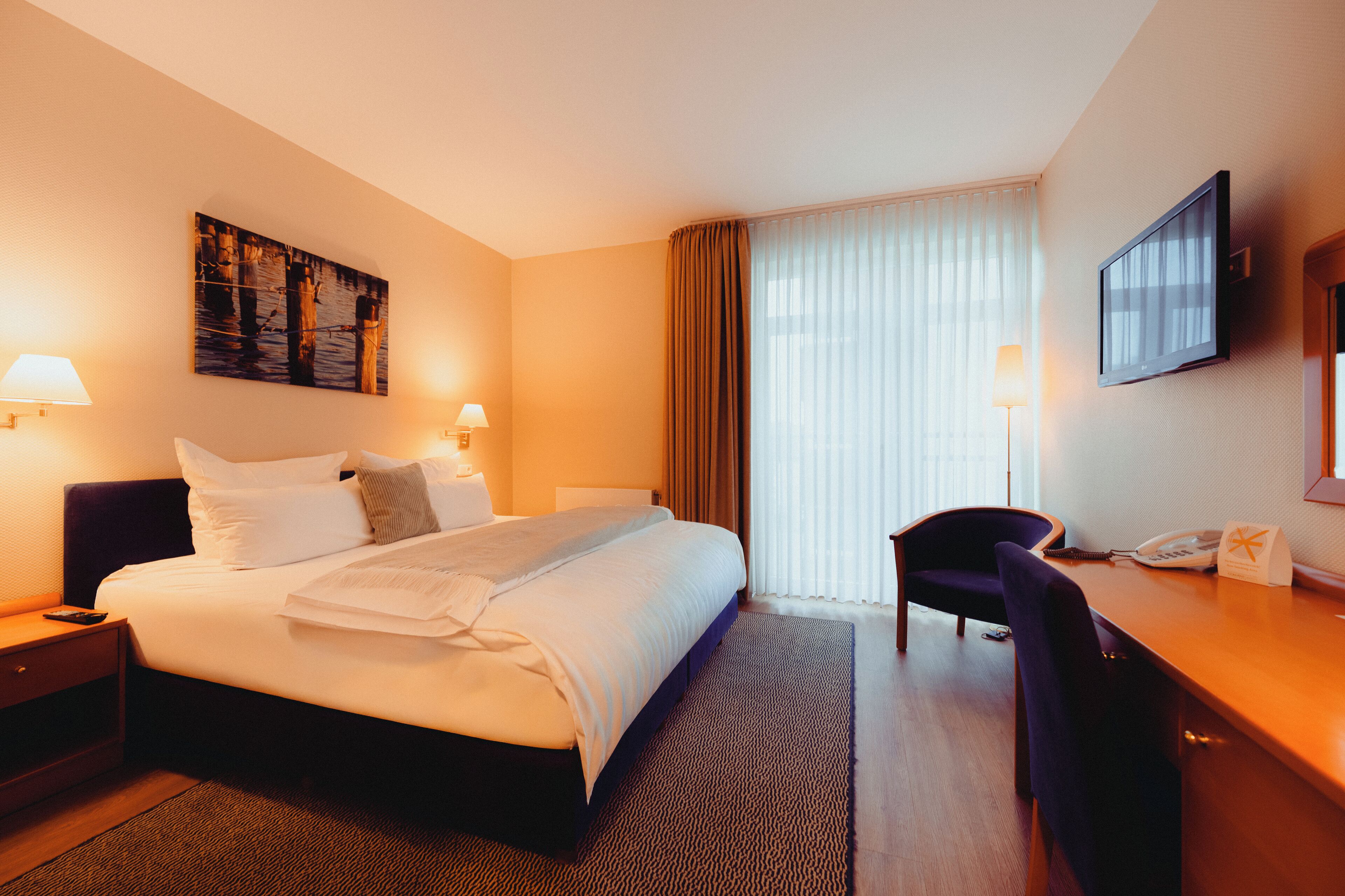 A hotel room with a large bed, two bedside tables with lamps, a desk with a chair, a flat-screen TV, and a painting on the wall.