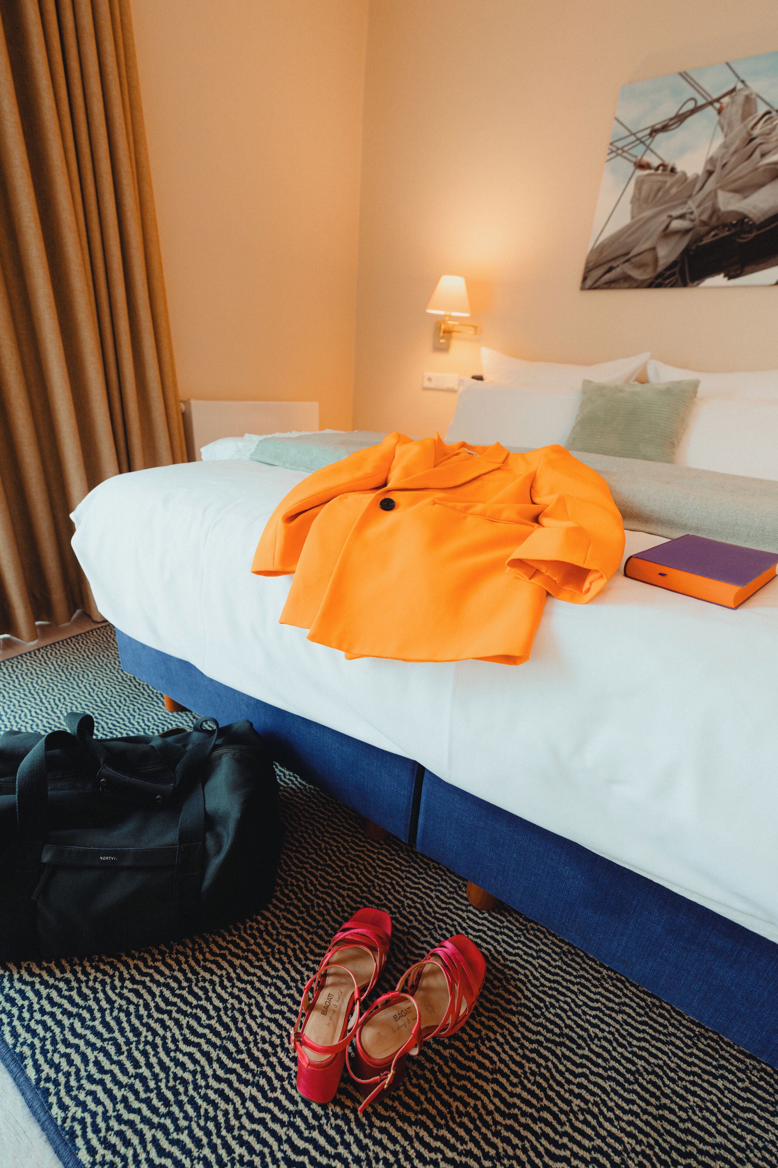 Un lit bien fait, recouvert d’une couverture blanche et bleue, une veste orange, une paire de chaussures rouges et un sac noir posé sur le sol.