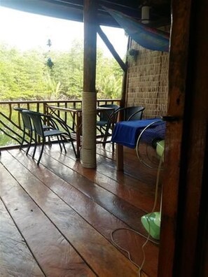 A covered outdoor area with wooden flooring, metal chairs, a table with a blue tablecloth, and a green fan. - Mangroves & More (Sihanoukville)