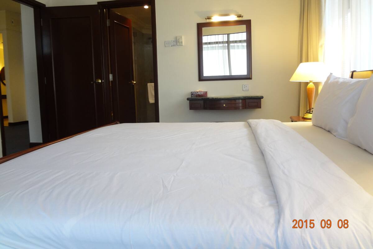 Una habitación de hotel con una cama grande, un espejo sobre el tocador, una mesita de noche con una lámpara y una puerta que da a otra habitación.