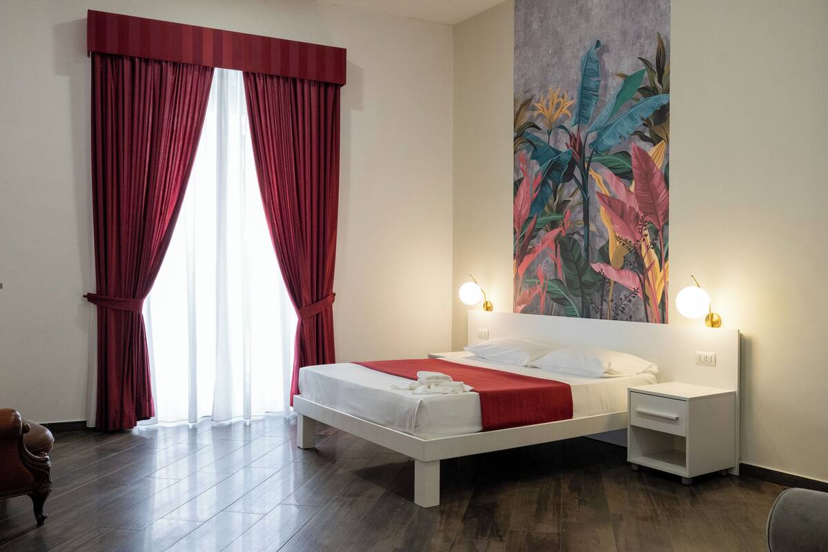 A hotel room with a large bed, a nightstand, a wall-mounted lamp, and a window with red curtains.
